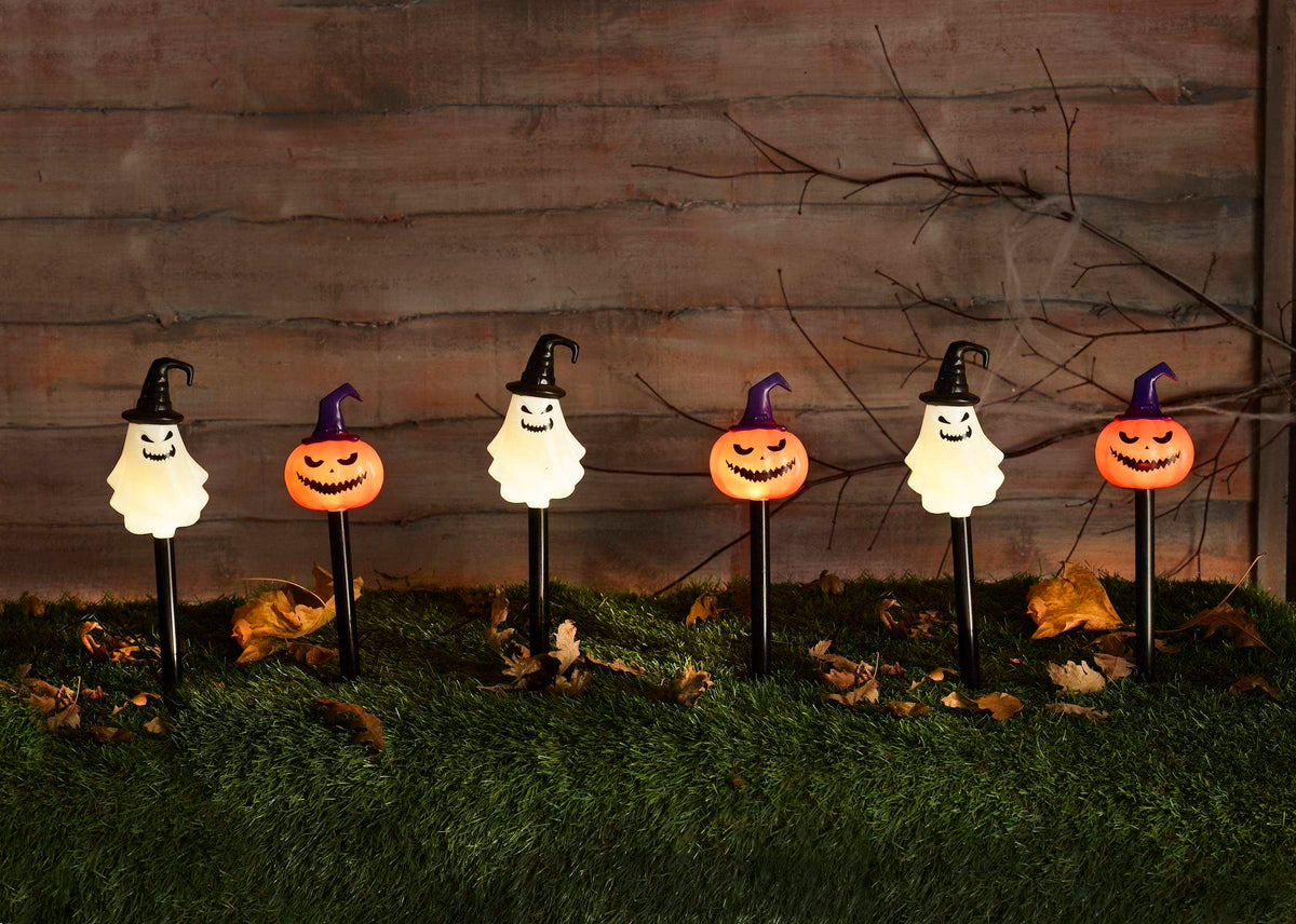 Six Halloween yard lights arranged in a row on artificial grass against a rustic wooden wall. The lights include four white ghost shapes with black witch hats and two orange pumpkin jack-o'-lantern faces with purple hats.