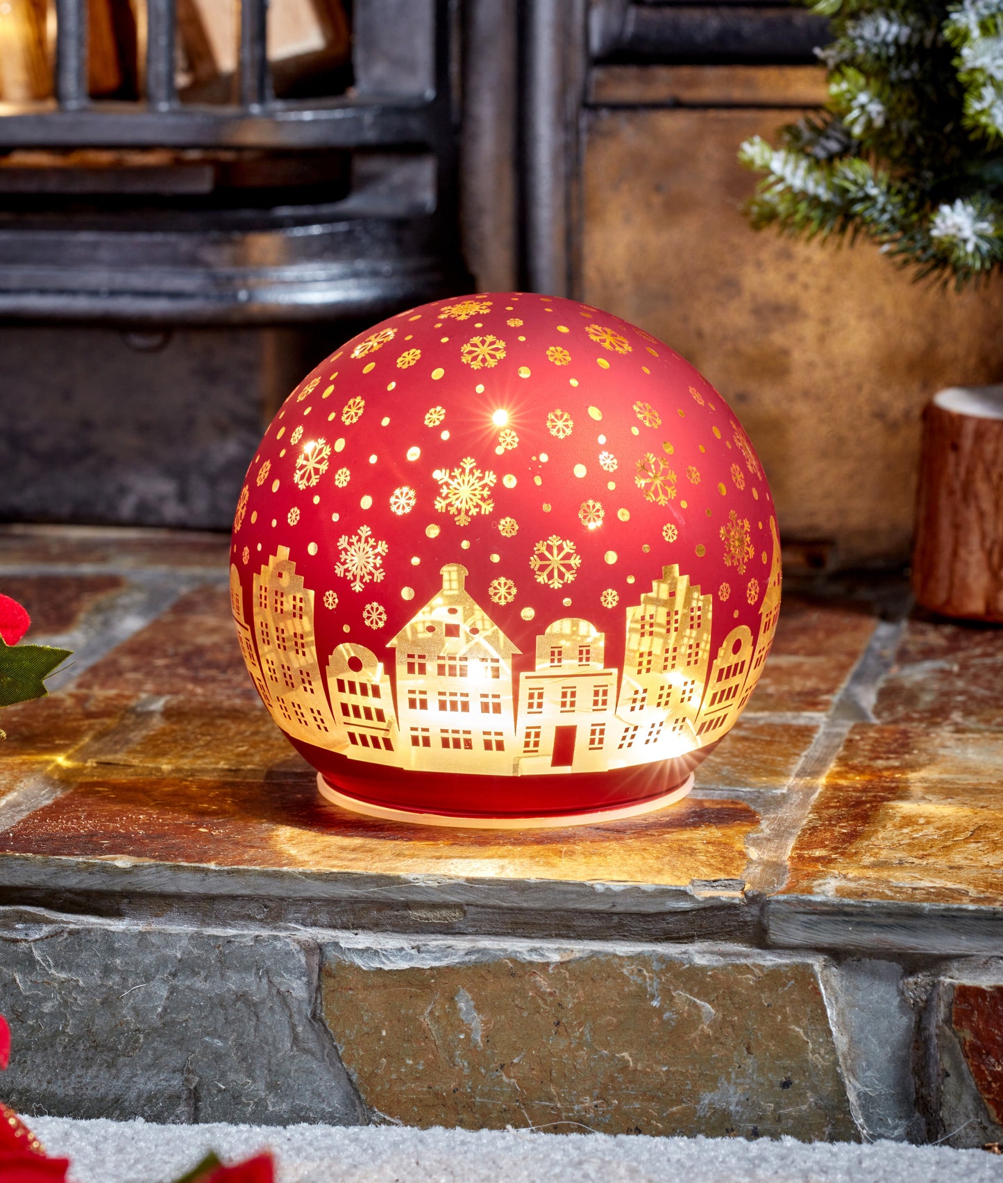 Decorative red lamp with gold cityscape design on a stone surface with a fireplace and Christmas tree in the background.