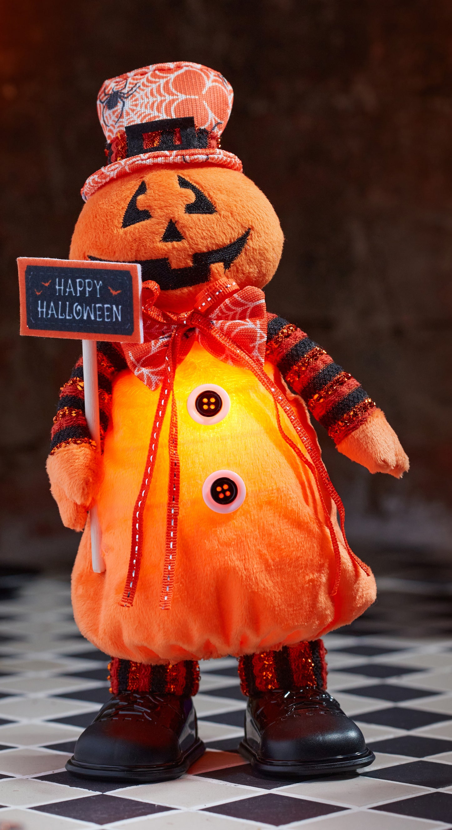 Halloween-themed plush pumpkin with a sign saying 'Happy Halloween' on a checkered floor.