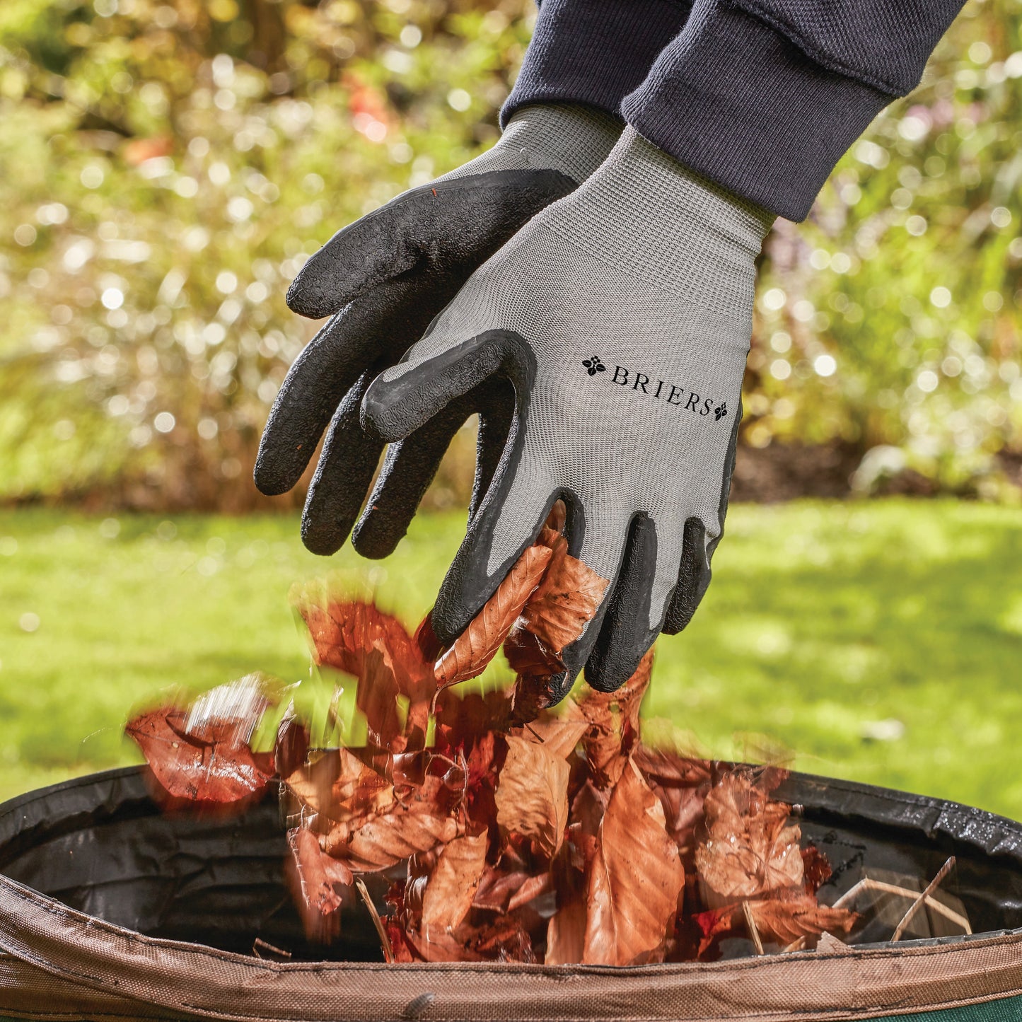 Person wearing Briers gloves collecting leaves in a garden bin with a blurred garden background