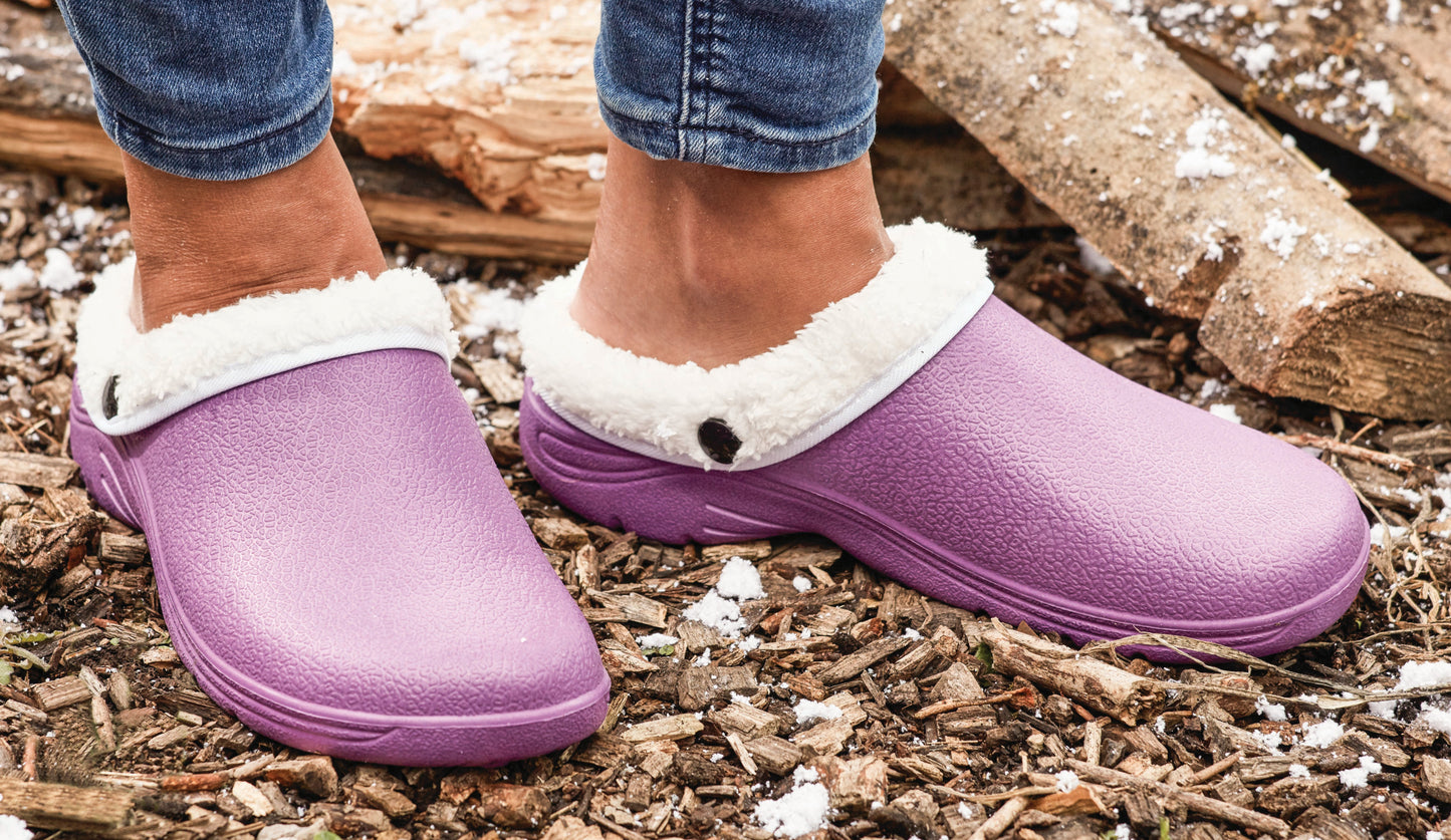Purple clogs with white fur trim worn by a person on a wooden floor.