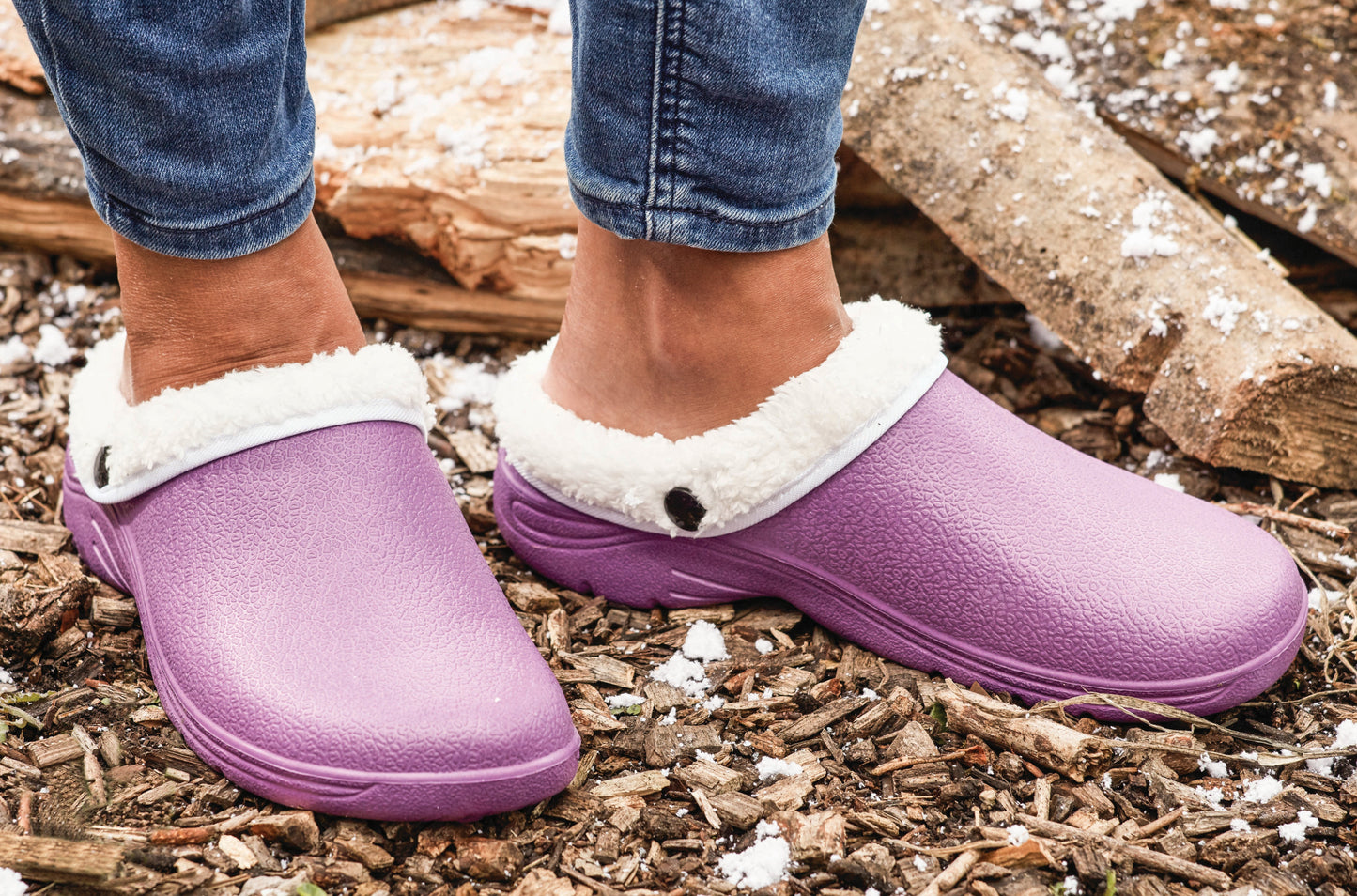Purple clogs with white fur trim worn by a person on a wooden ground.