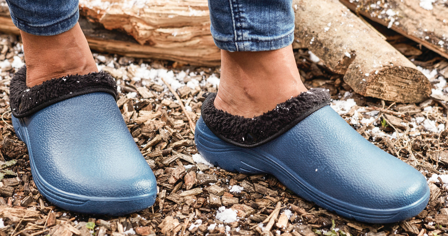 Blue clogs with fuzzy black trim worn by a person on a wooden floor.