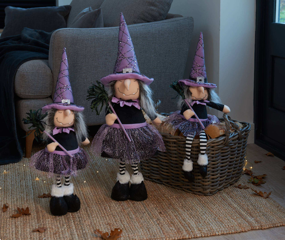 Three witch-themed plush dolls with purple pointed hats, black and white striped outfits, and gray hair, arranged on a woven rug in front of a gray sofa. One doll sits in a wicker basket, and small string lights and scattered autumn leaves decorate the floor.