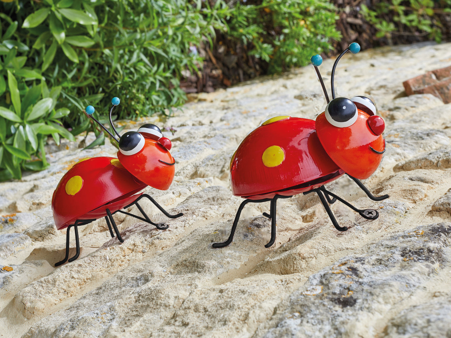 Two red ladybug figurines on a stone surface with greenery in the background