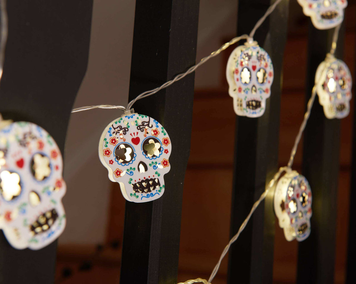 Dia de los Muertos sugar skull string lights hanging in front of black metal railings, featuring white skulls decorated with colorful floral patterns and rhinestone eyes.