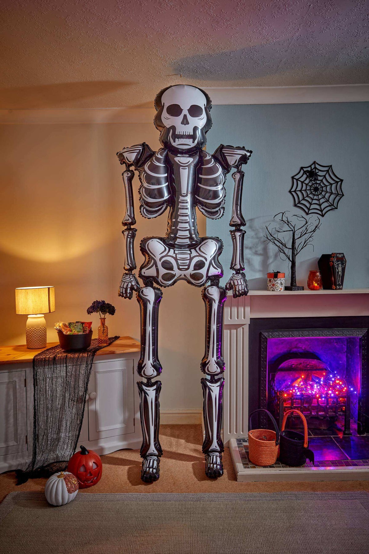 A large inflatable skeleton Halloween decoration stands in a living room in front of a fireplace. The room is decorated for Halloween with a lit fireplace, spider web wall decor, a small pumpkin, and a black net on the floor.