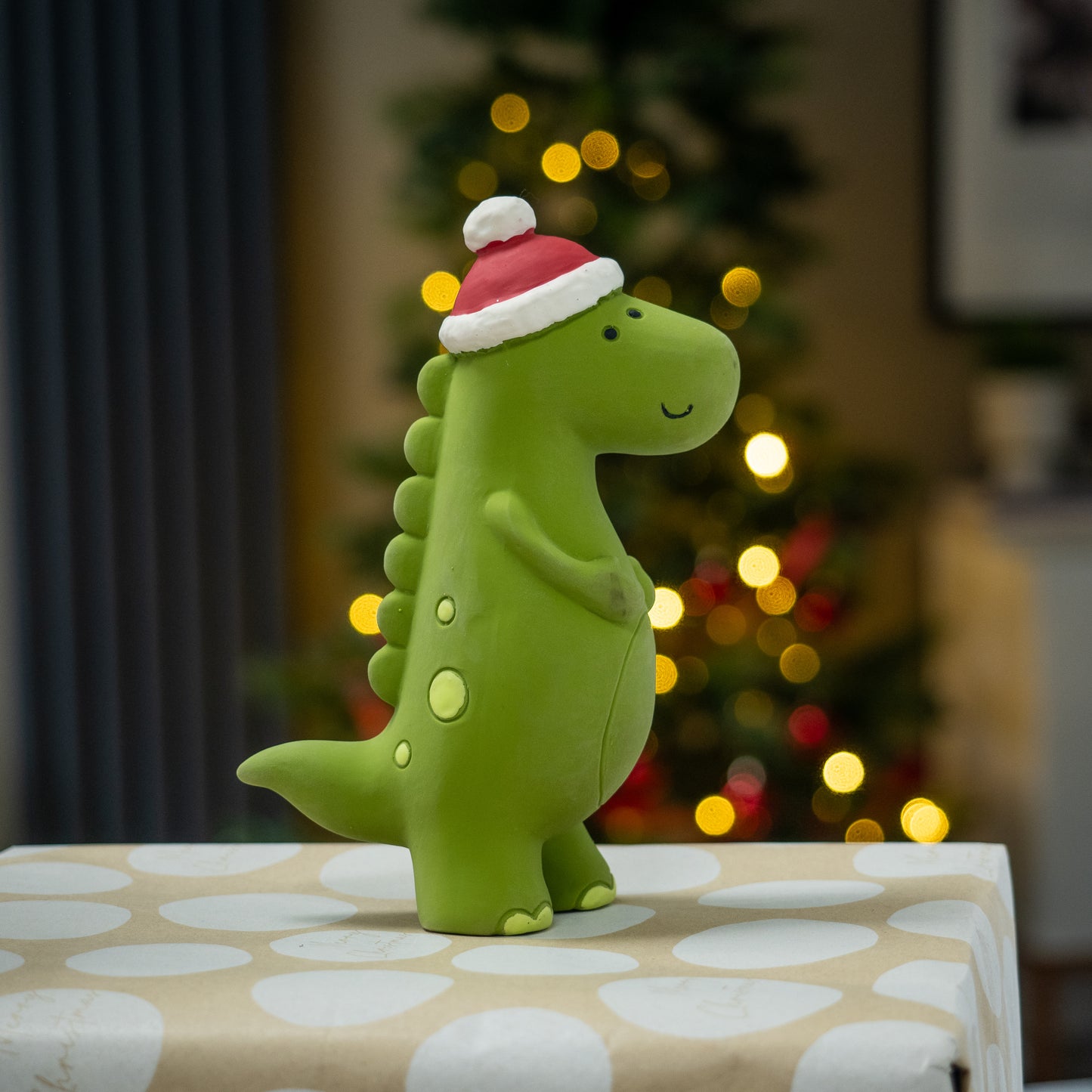Green dinosaur toy with a Santa hat on a wrapped gift with a blurred Christmas tree in the background.