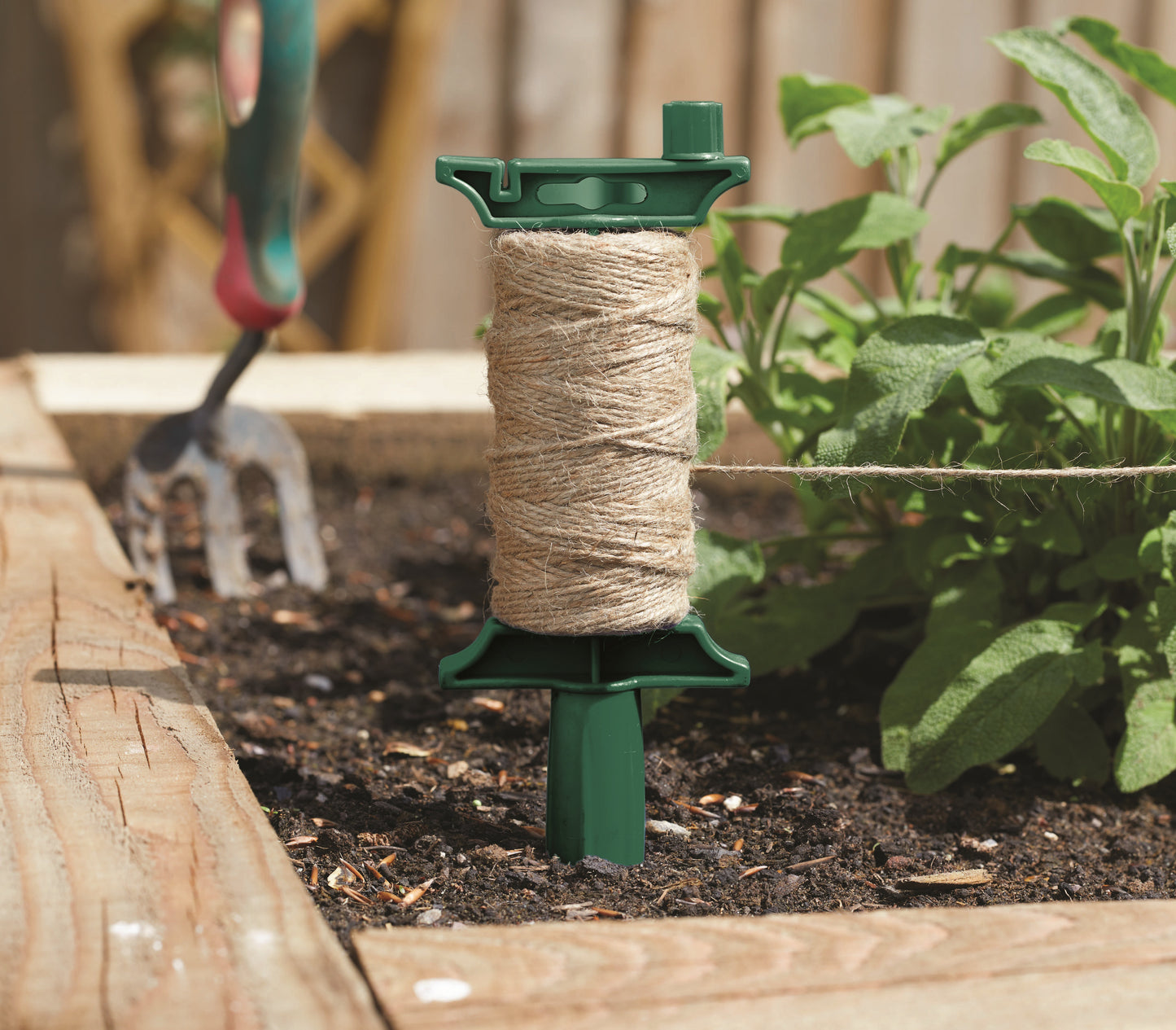 Spool of twine on a green stand in a garden setting with plants and gardening tools.