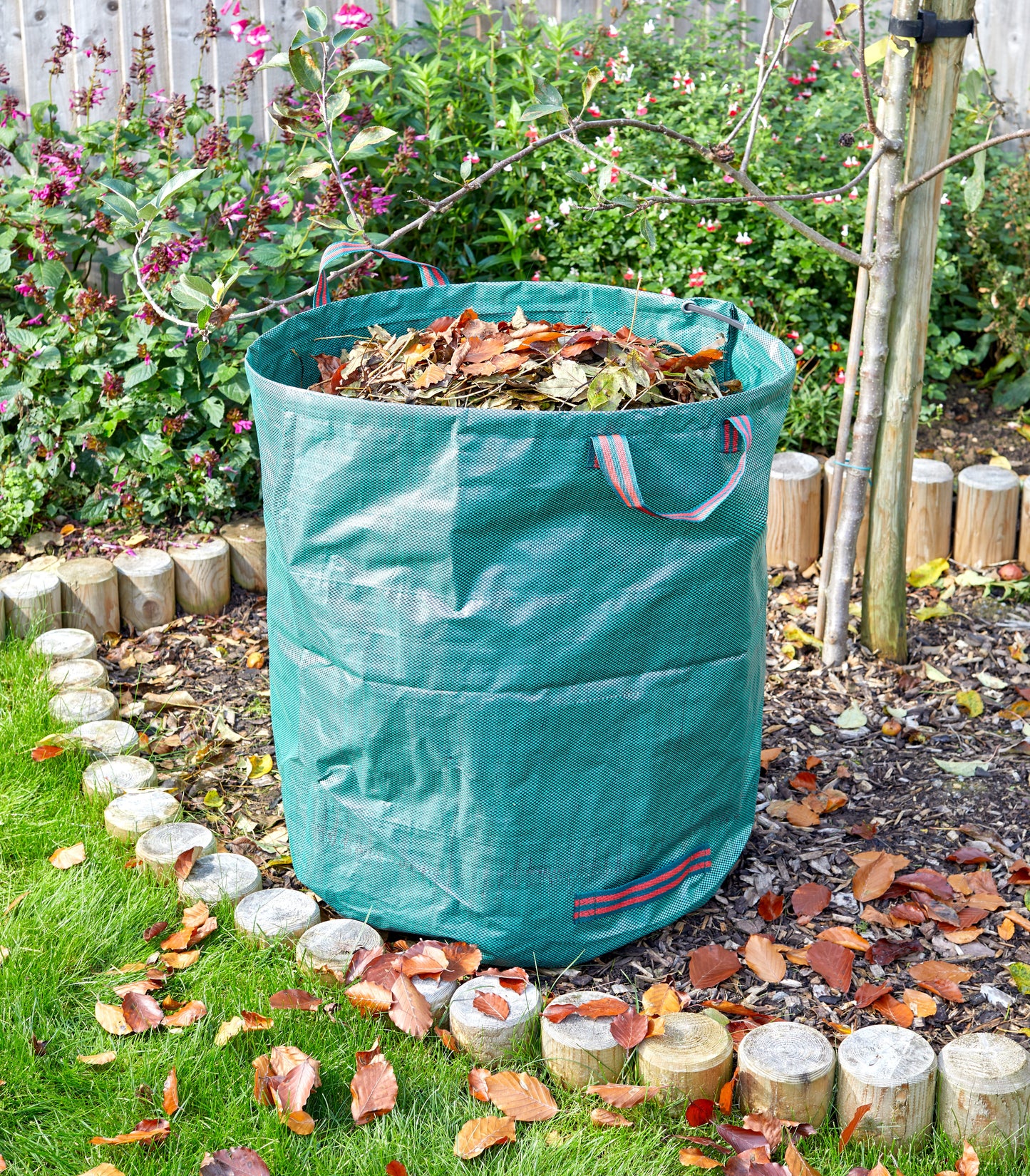 Green garden waste bag filled with leaves in a garden setting.