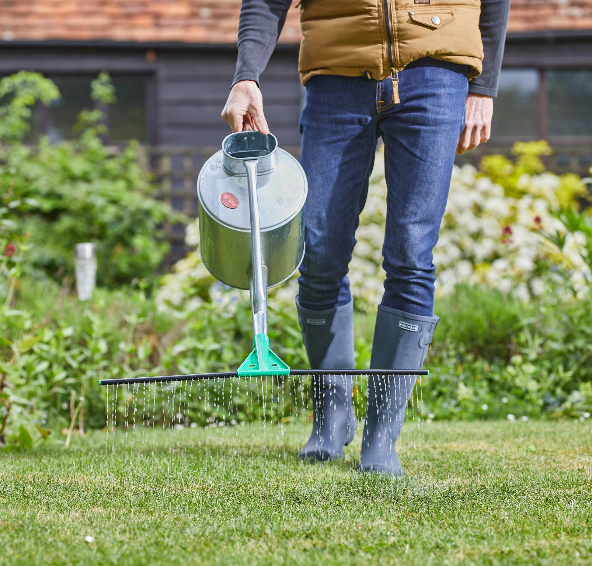 Person using a garden sprinkler on a grassy area with a garden and house in the background