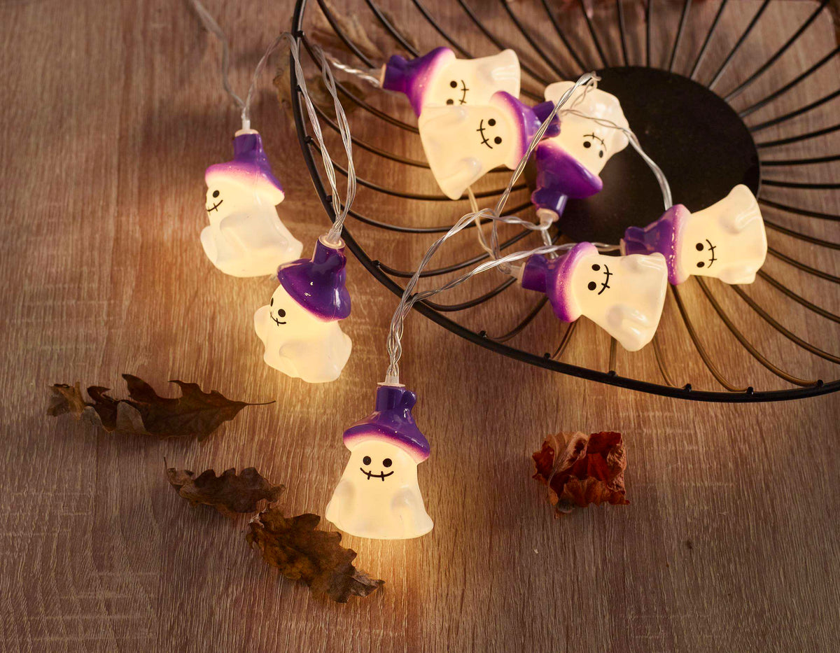 Halloween ghost string lights with glowing white bodies and purple wizard hats, displayed on a wooden surface next to a black wire basket.