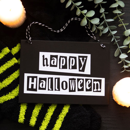 Black sign with 'happy Halloween' text on a dark background with greenery and candles.