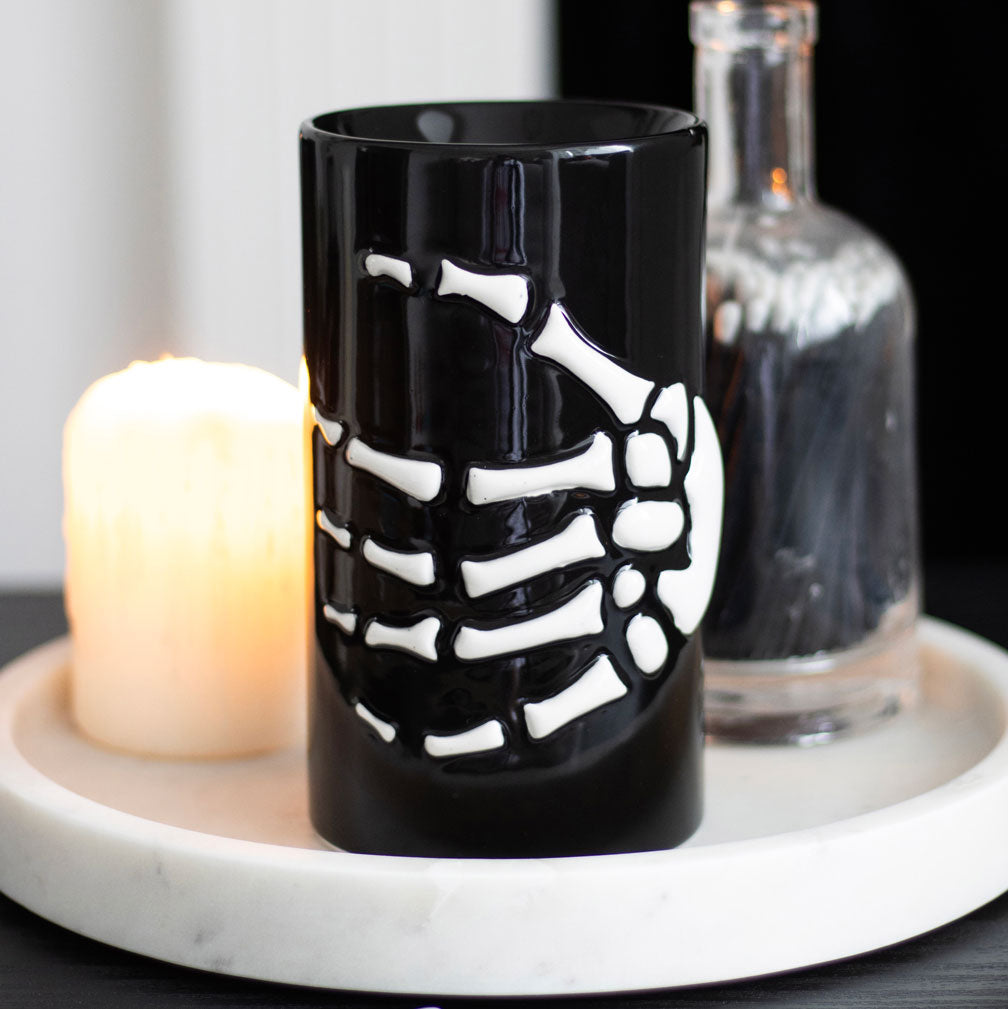Black mug with skeleton design on a white surface with a candle and glass bottle in the background.