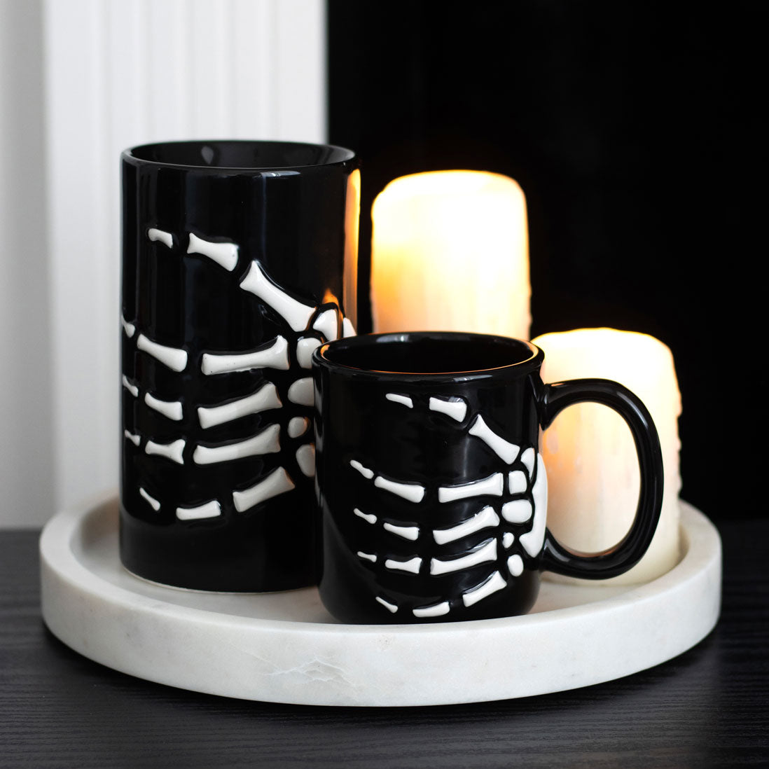 Black skeleton-hand and bone-patterned mugs on a marble tray with candles in the background.