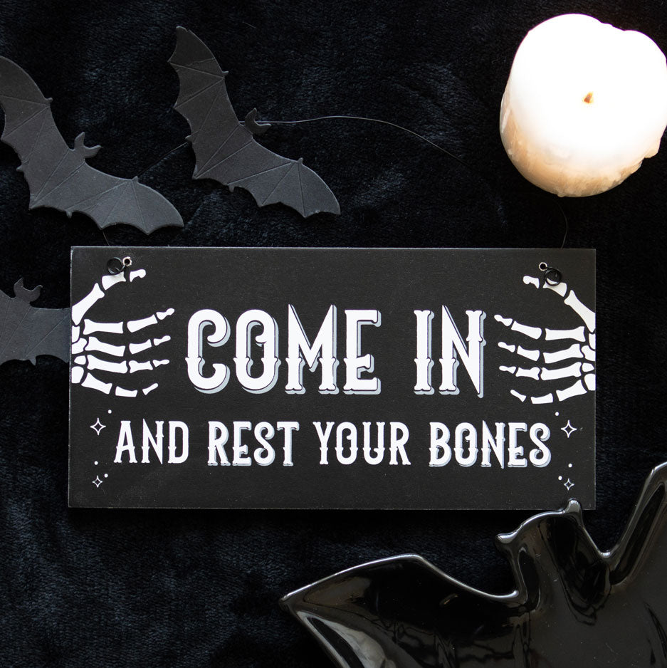 Halloween-themed sign with skeleton hands and text 'Come in and rest your bones' on a dark background with bats and a candle.
