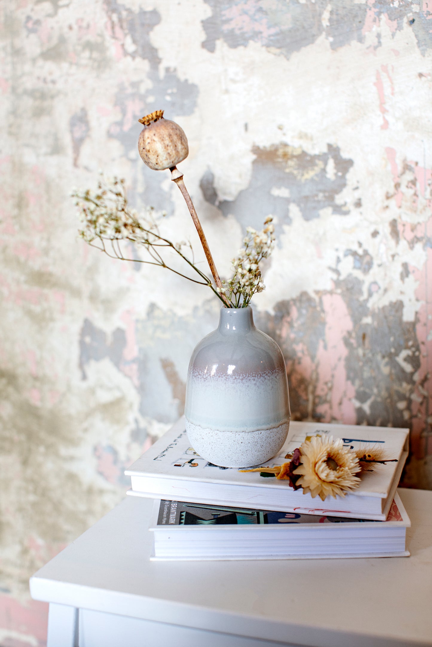 White vase with dried flowers on top of books against a textured wall.