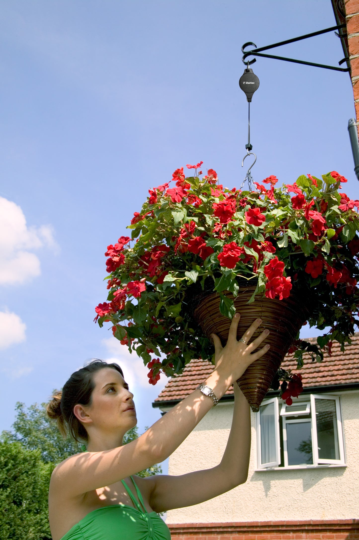 Woman adjusting a hanging basket of red flowers against a clear blue sky.