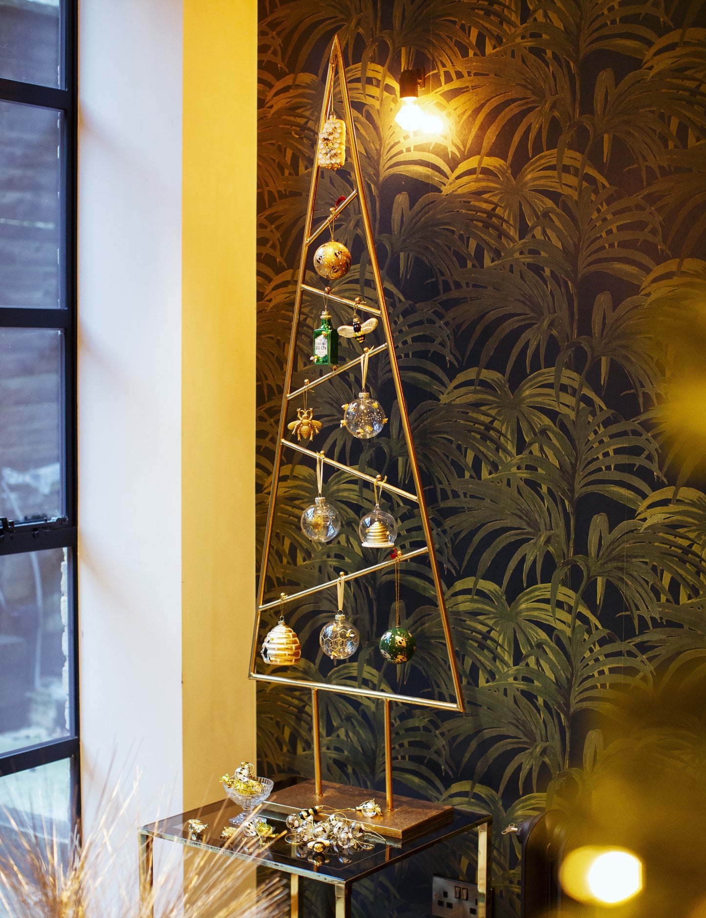 Decorative Christmas tree with ornaments on a stand against a leafy wallpapered wall.