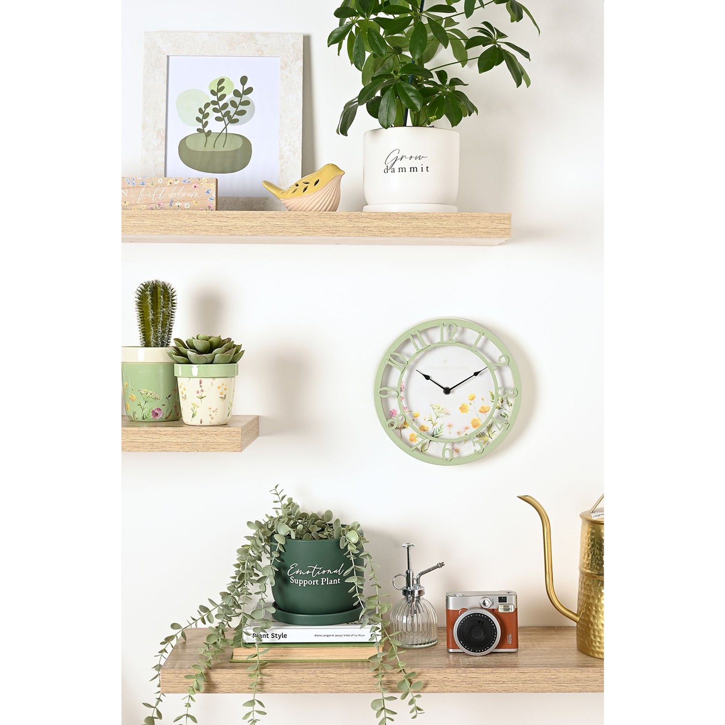 Decorative wall clock with plants and books on a shelf against a white wall.