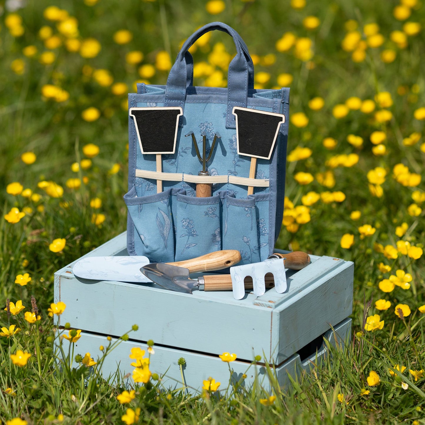 Blue gardening tool set with tools and bags on a wooden crate in a field of yellow flowers.