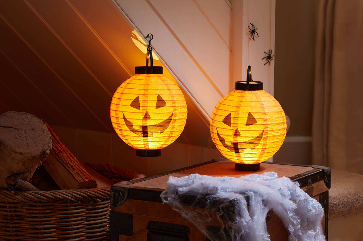 A pair of orange paper lanterns shaped like a jack-o'-lantern with a black hanging hook at the top.