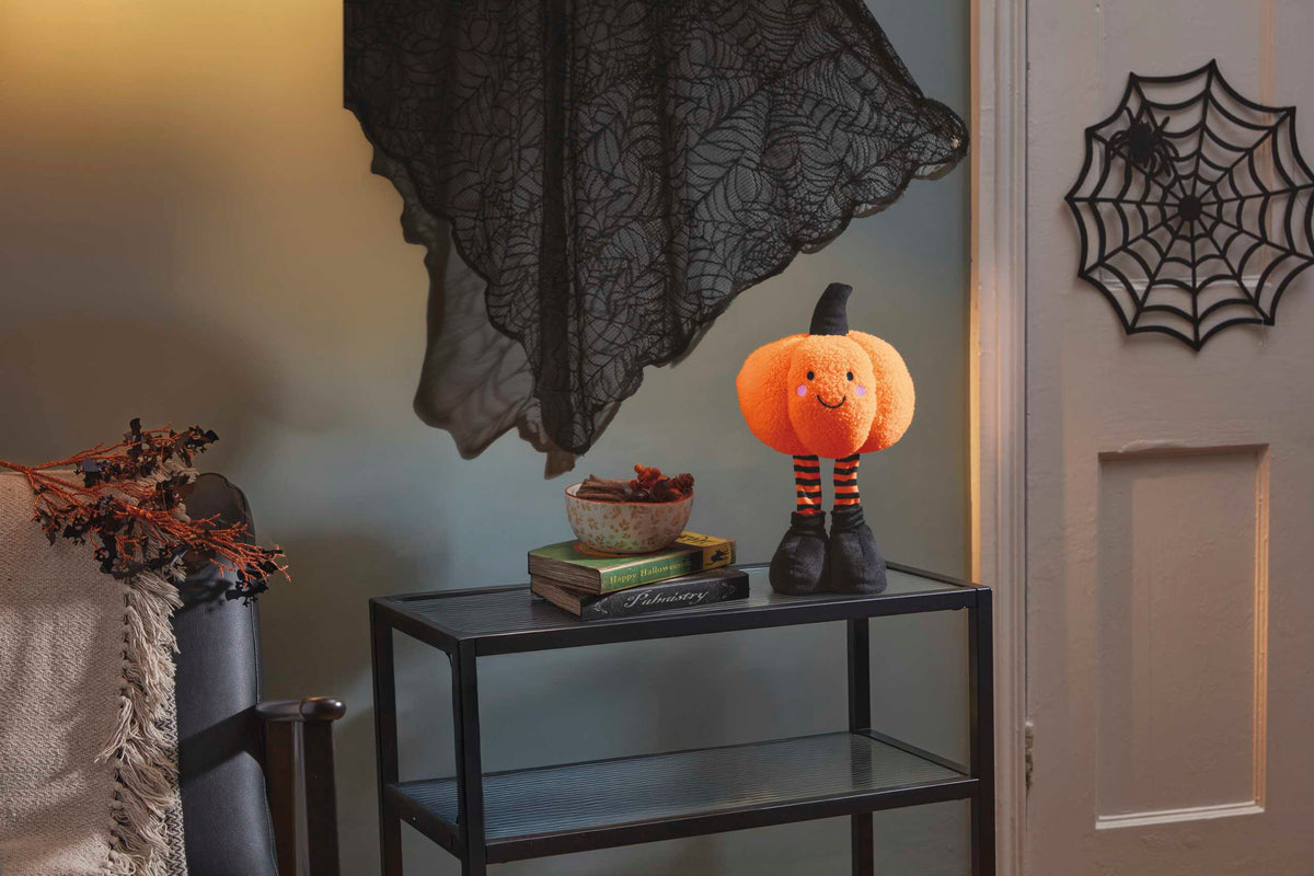A Halloween-themed orange pumpkin decoration with a stitched face and striped legs, standing on a small black side table in a styled room with a black lace curtain and spider decoration on the door.