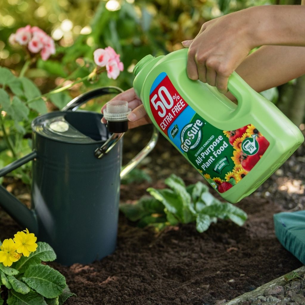 Person holding a green bottle of plant food with flowers and plants in the background