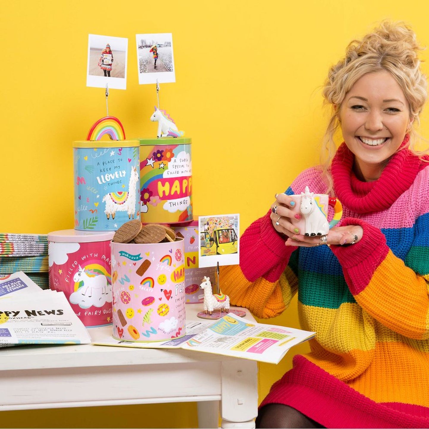 Woman in colorful sweater holding a unicorn-themed mug with decorative tins and stationery on a yellow background