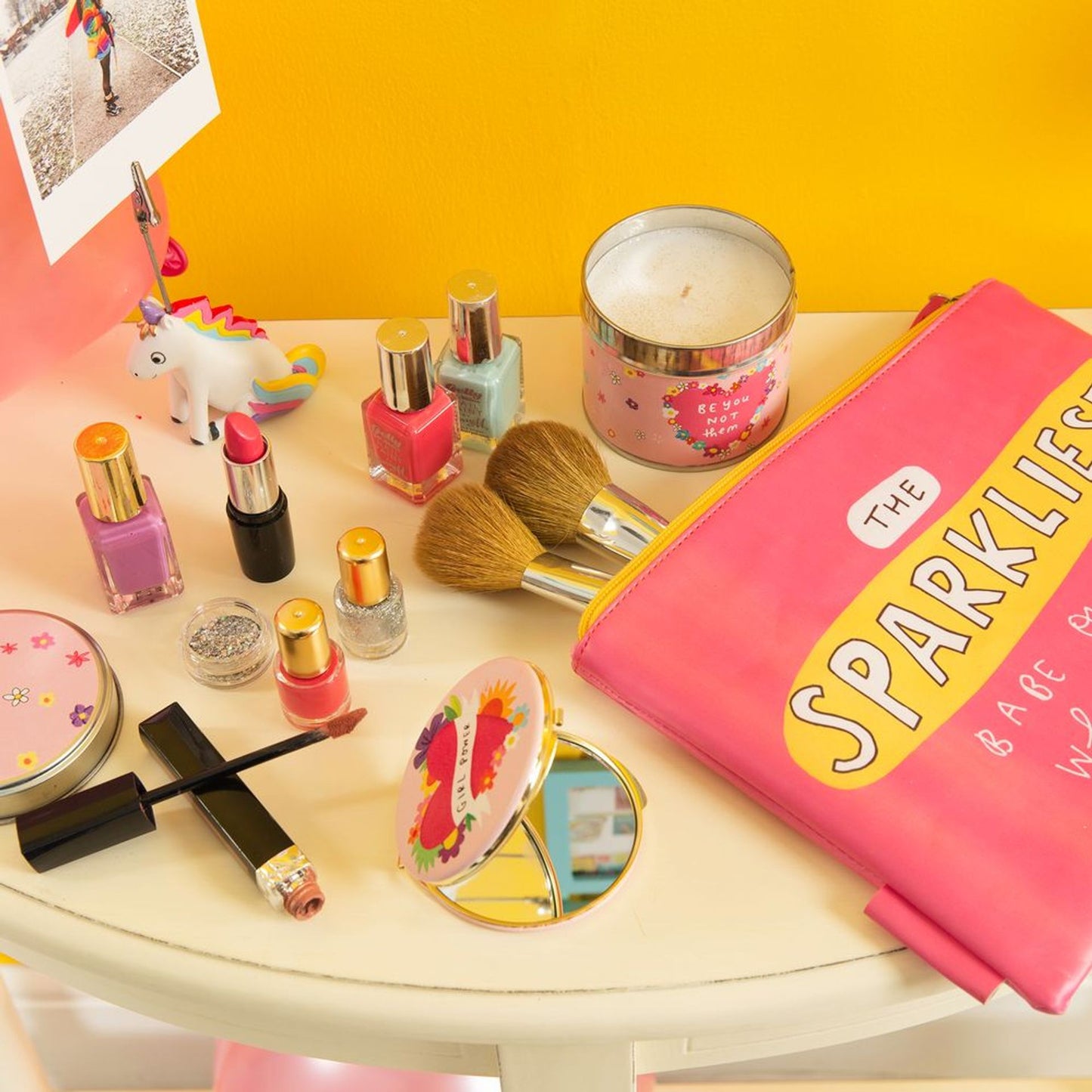 Makeup items including nail polishes, lipsticks, and a candle on a table with a pink pouch labeled 'The Sparklies' against a yellow background.