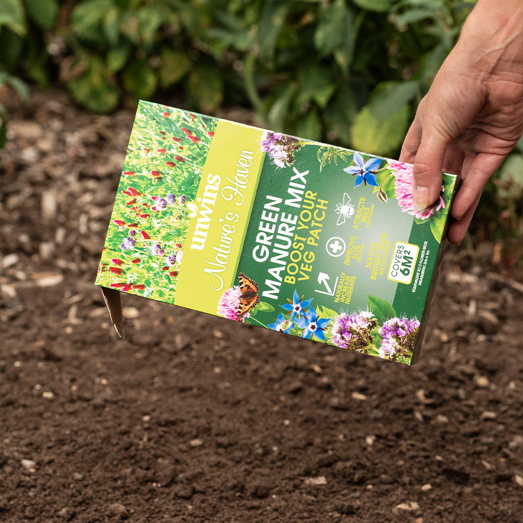 Hand holding a packet of Nature's Garden Green Manure Mix over garden soil.