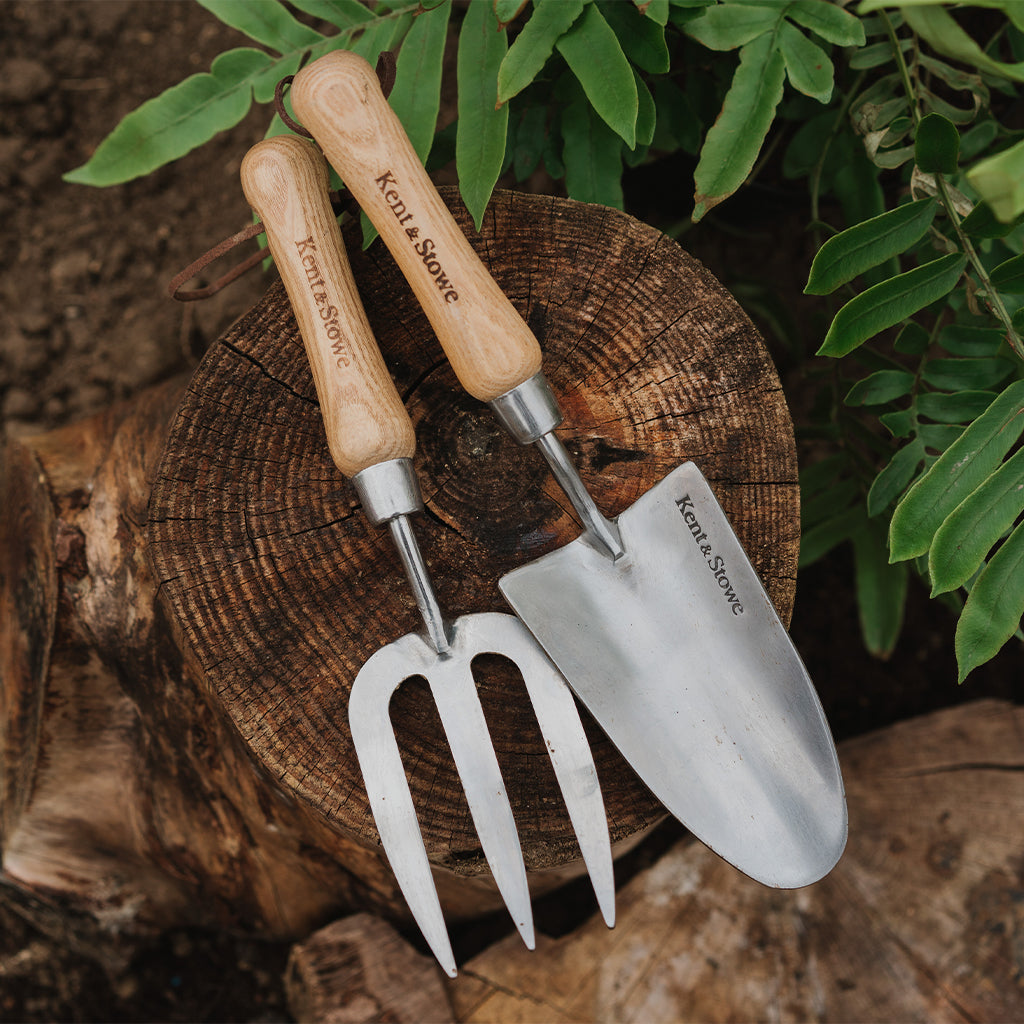 Two gardening tools, a fork and a trowel, on a wooden log with green foliage in the background.
