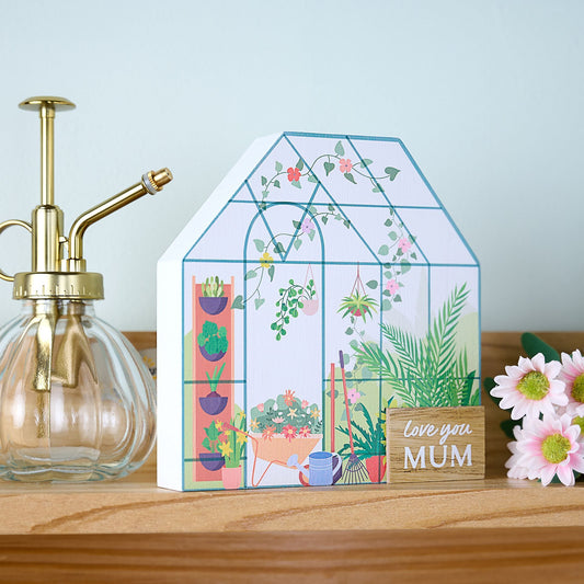 Decorative house-shaped card with plants and 'Love you MUM' text on a wooden surface with a spray bottle and flowers.