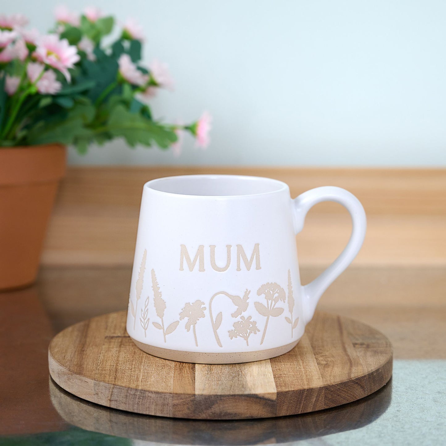 White mug with 'MUM' and floral designs on a wooden coaster, with a blurred background of flowers and a plant.