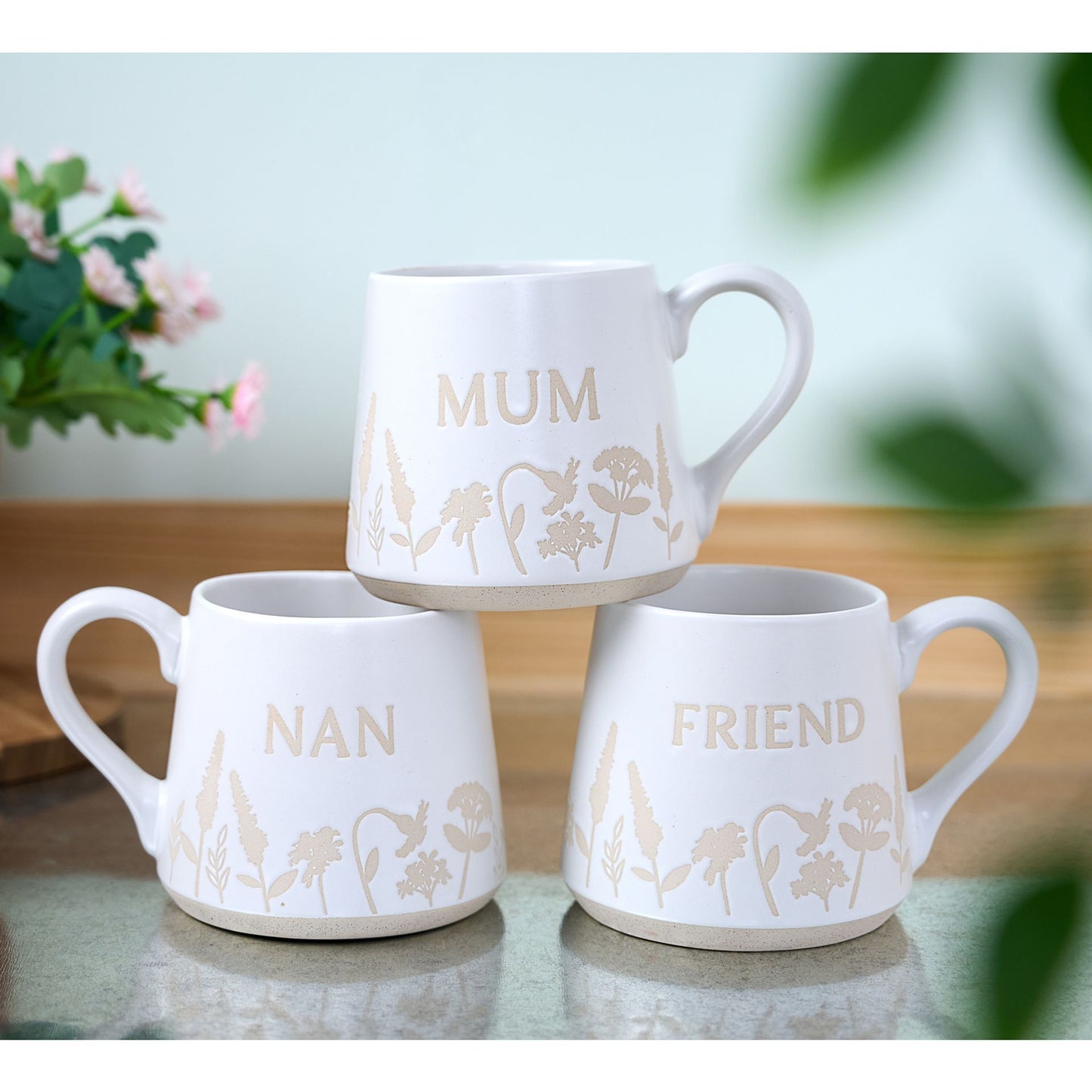 Three white mugs with floral designs and text on a table with a blurred background