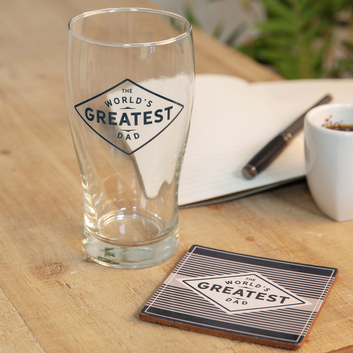 Clear glass with 'The World's Greatest Dad' design on a wooden table with a coaster.