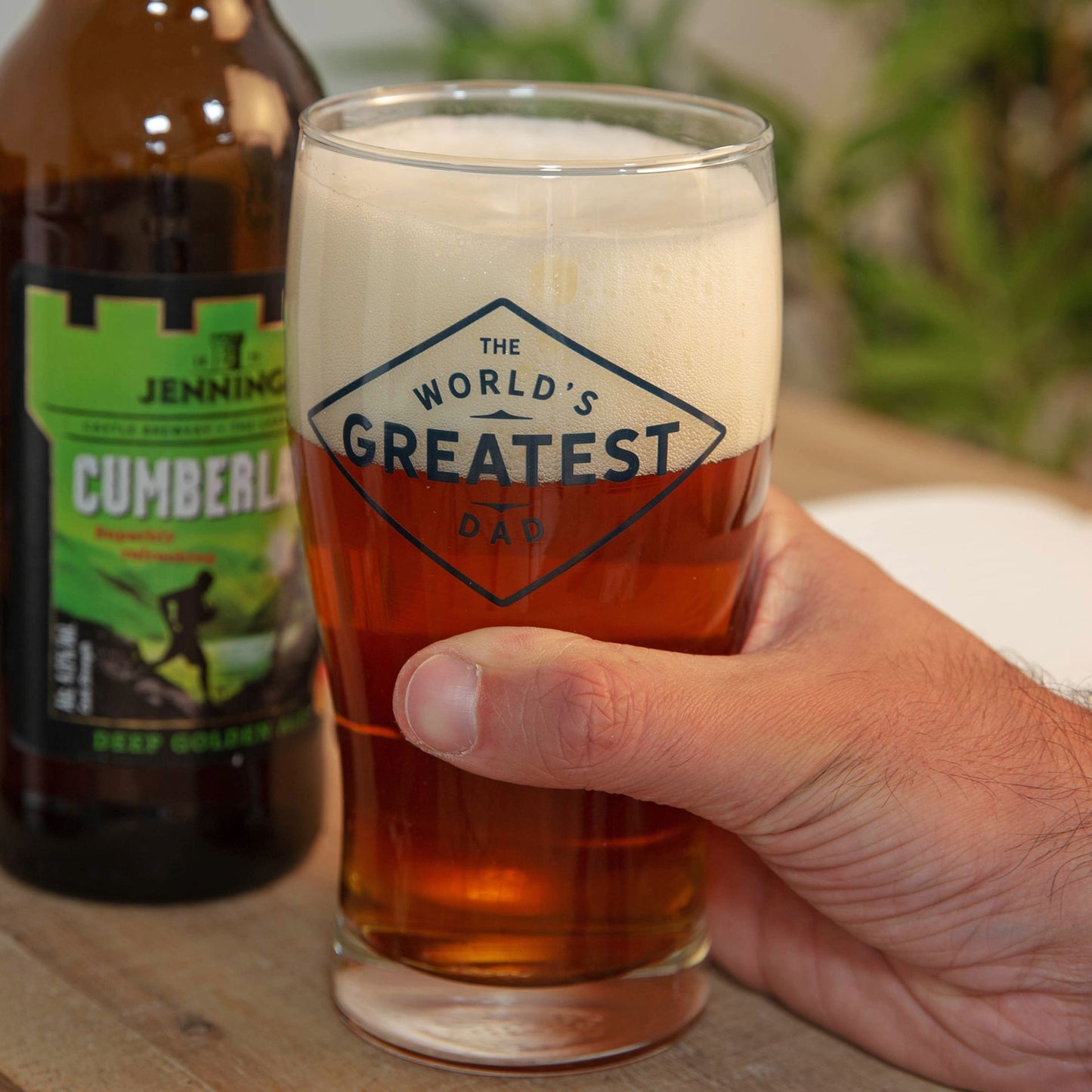 Hand holding a glass of beer with 'The World's Greatest Dad' text, next to a bottle labeled 'Cumberland'.