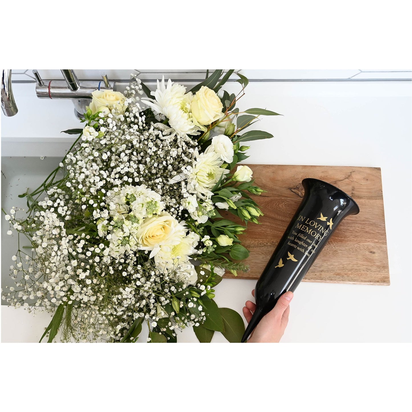Bouquet of flowers on a wooden board with a black urn labeled 'In Loving Memory' held by a hand.