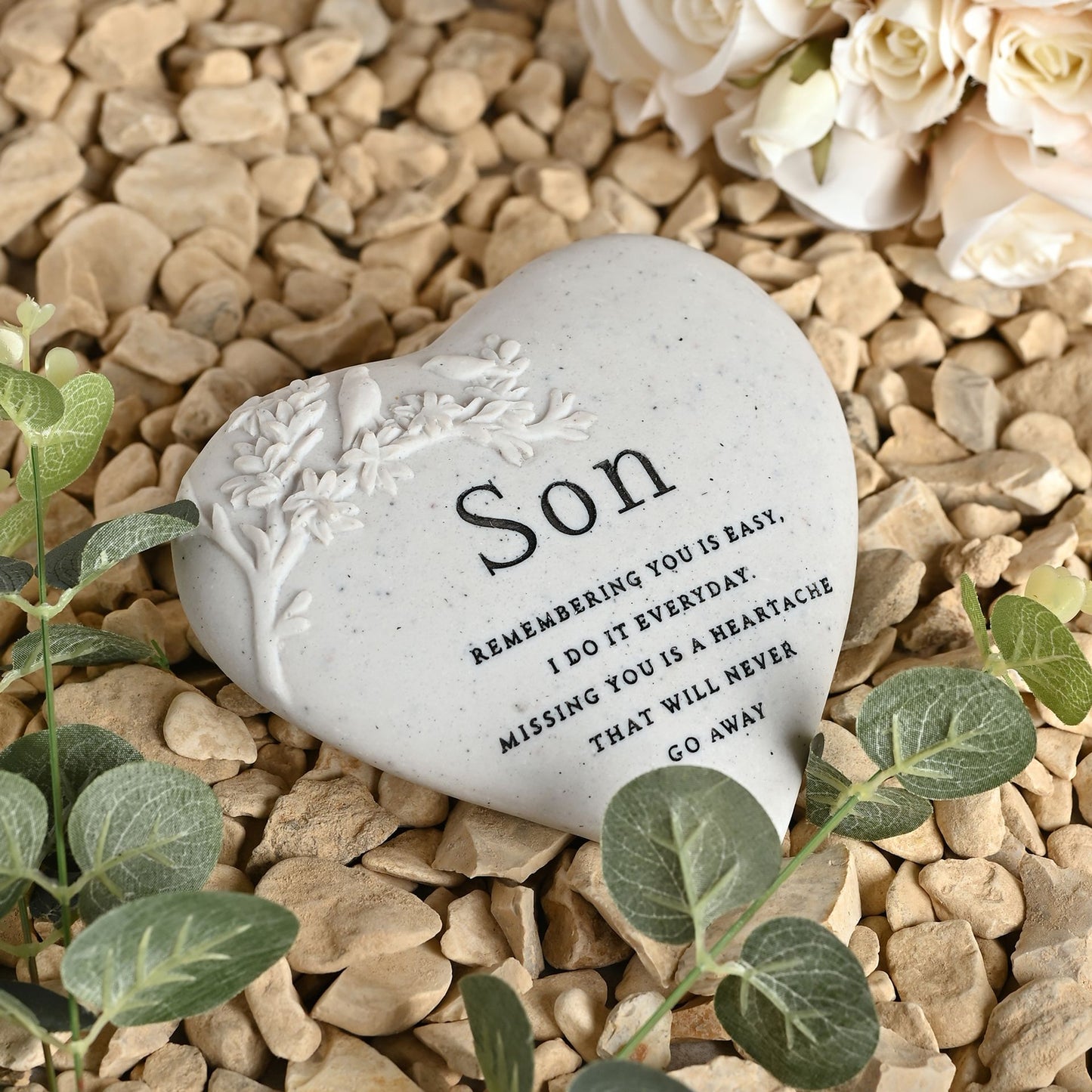 Heart-shaped stone with engraved text on a pebble surface with flowers and leaves.