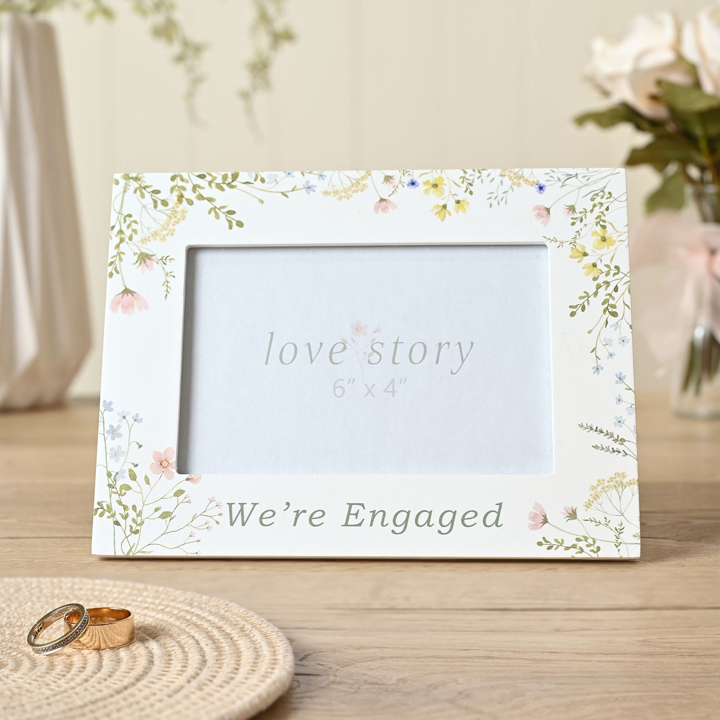 Floral photo frame with 'love story' and 'We're Engaged' text on a wooden surface.