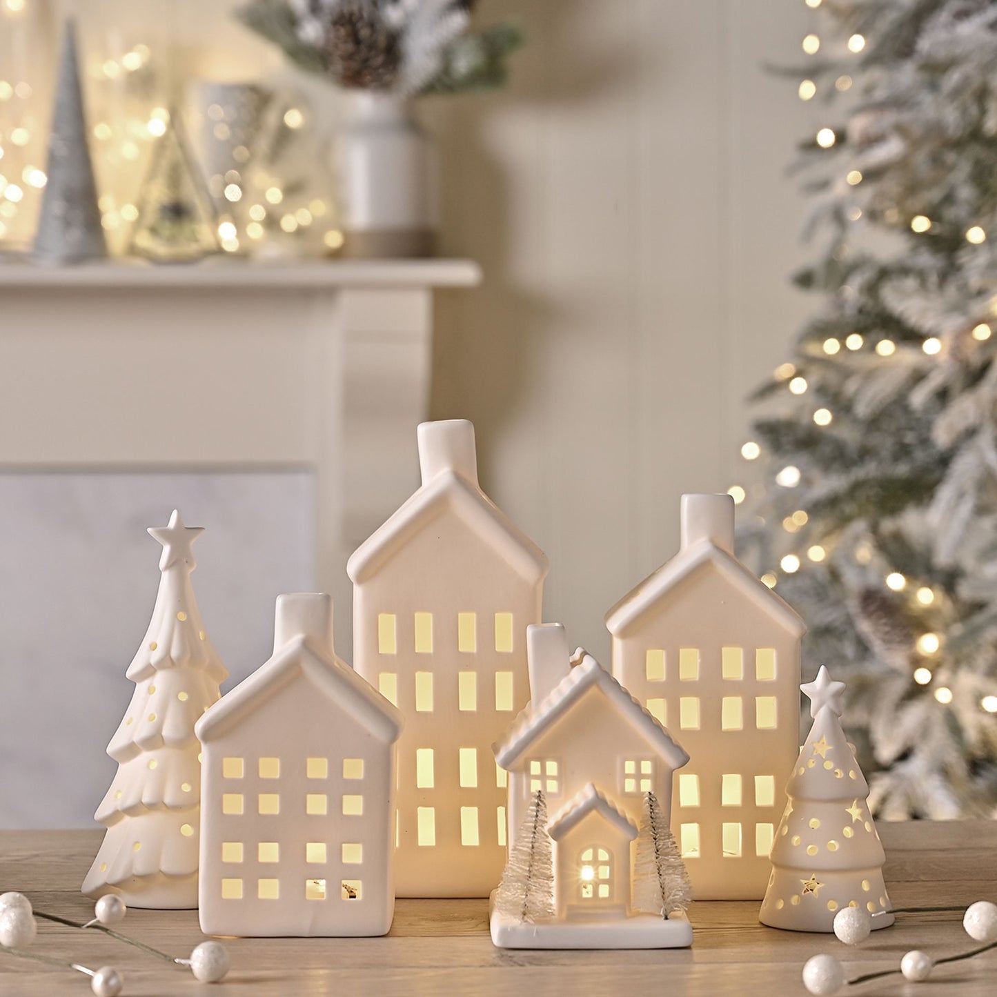 Set of ceramic houses with lit interiors on a wooden surface, Christmas tree and fireplace in the background.