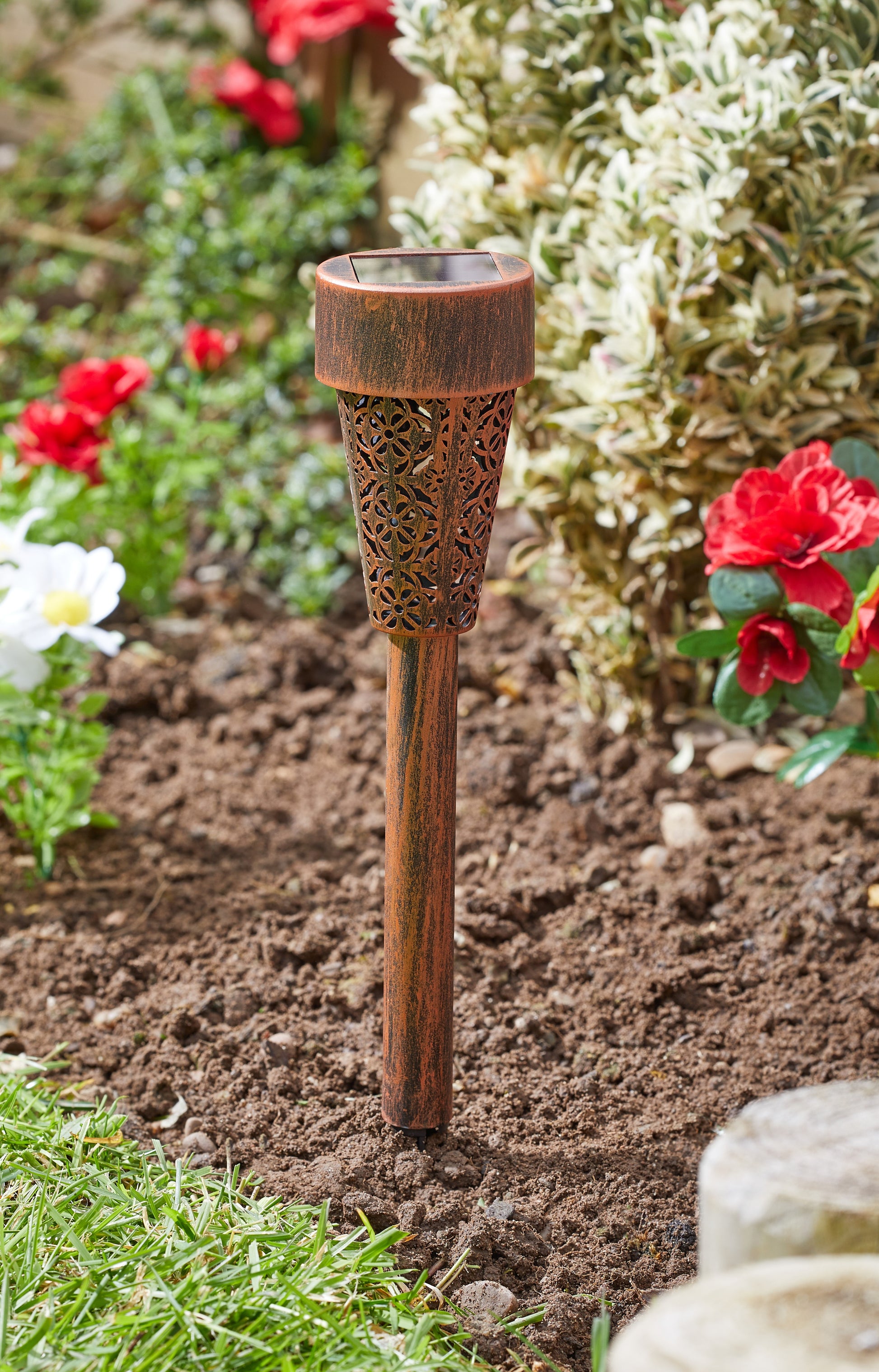 Decorative garden torch with intricate design in a garden setting with flowers and soil.