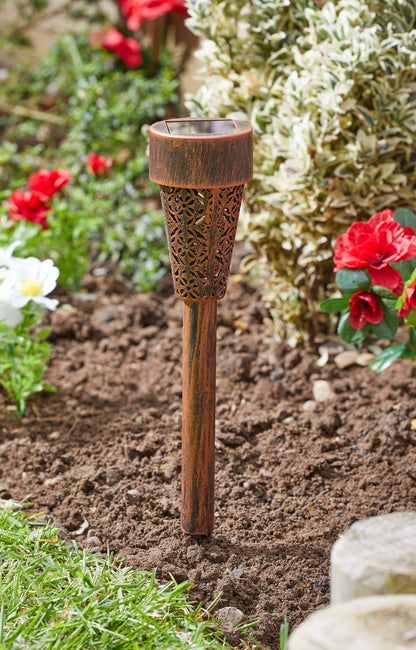Decorative garden torch with intricate design in a garden setting with flowers and soil.