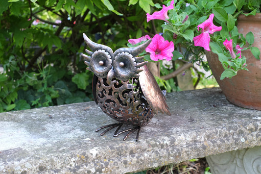 Decorative metal owl sculpture on a stone ledge with pink flowers in the background