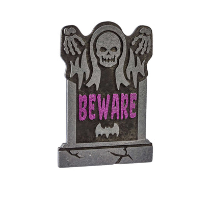 A Halloween tombstone decoration featuring a gray skull with wings, ghostly hands, and the word 'BEWARE' in pink glittery letters on a white background.