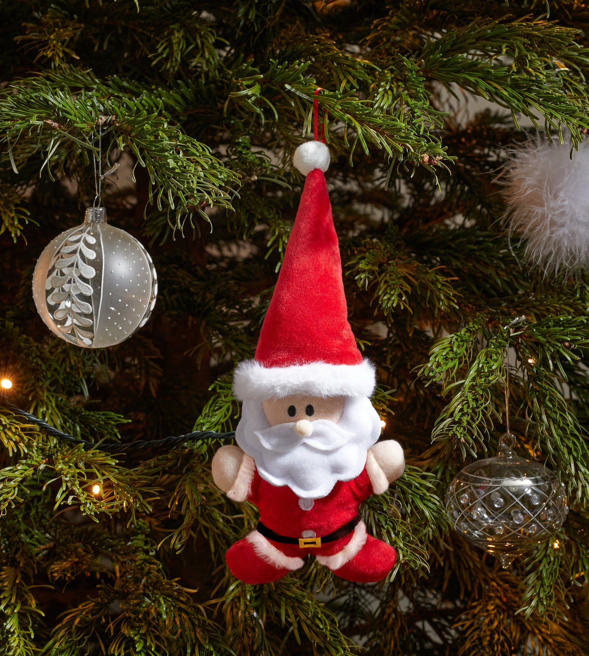 Plush Santa Claus ornament hanging on a decorated Christmas tree with glass ornaments.