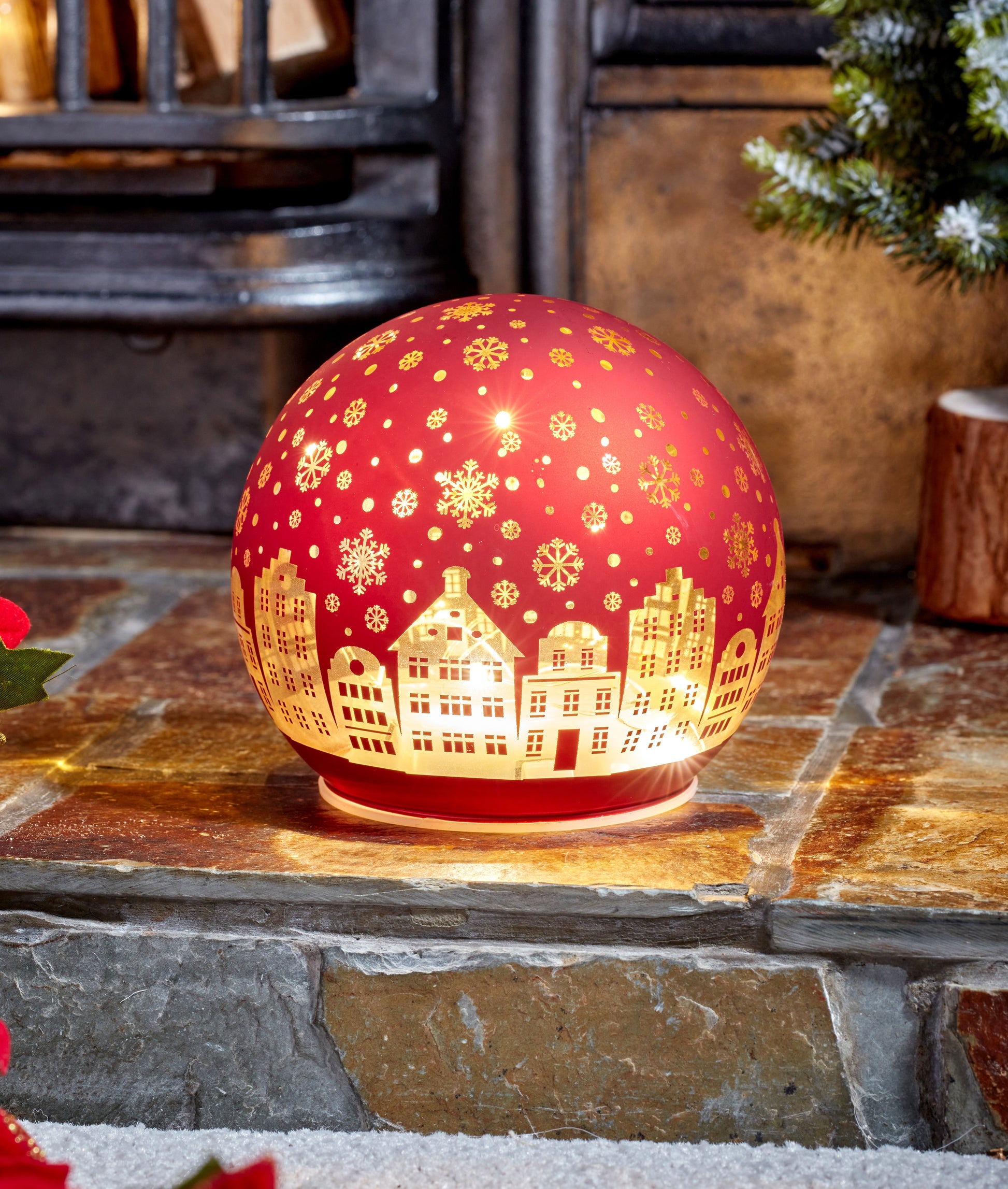 Decorative red lamp with gold cityscape design on a stone surface with a fireplace and Christmas tree in the background.