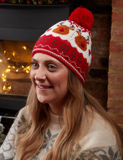 Woman wearing a red knit hat with reindeer design in front of a fireplace.