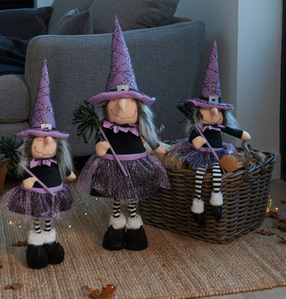 Three witch figurines with purple hats and dresses on a woven basket against a neutral background.