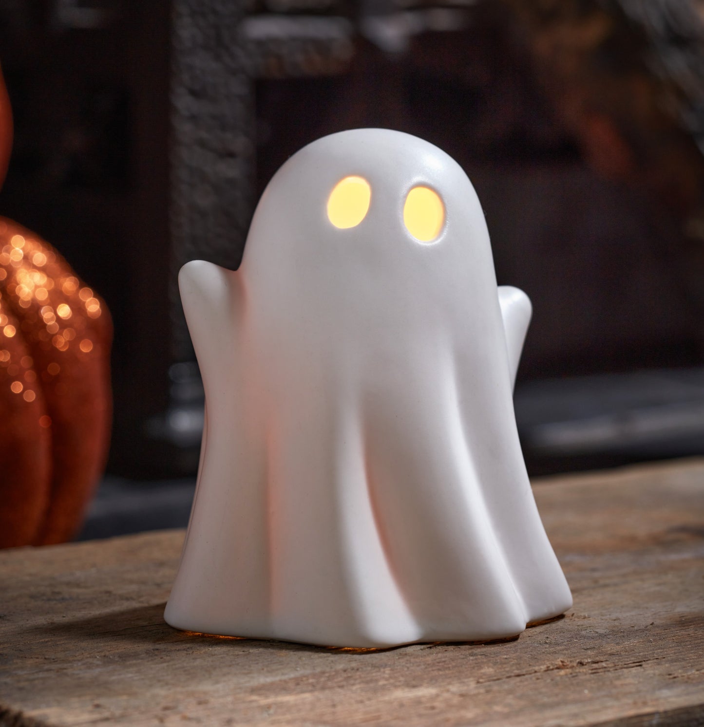 White ghost-shaped night light with glowing eyes on a wooden surface, blurred pumpkin in the background